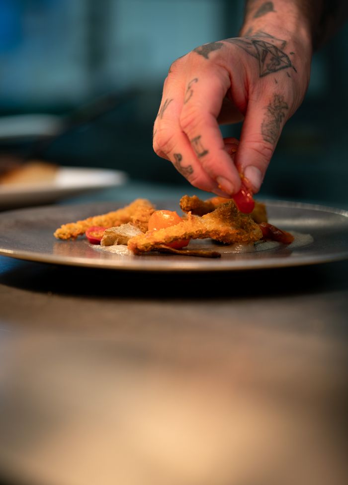 Close-up of a tattooed hand garnishing crispy food with cherry tomatoes on a plate in a kitchen setting.